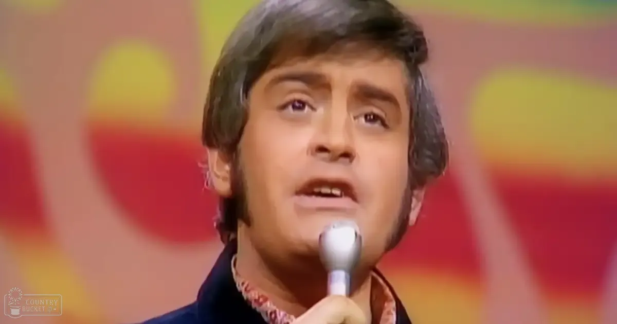 A middle-aged man singing into a microphone in what appears to be a studio setting with warm, vibrant background lighting and mid-20th century style clothing.