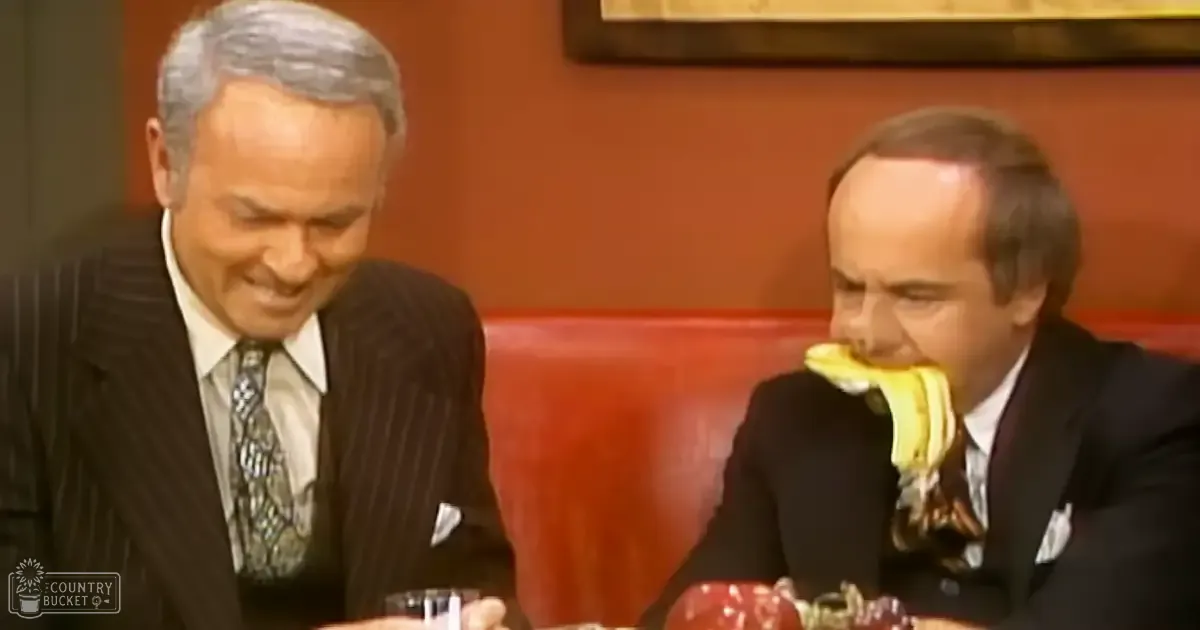 Middle-aged male in a dark pinstripe suit and patterned tie interacts with another middle-aged to older adult male holding a banana near his mouth, both seated at a red leather booth in an indoor setting suggestive of a restaurant or bar.
