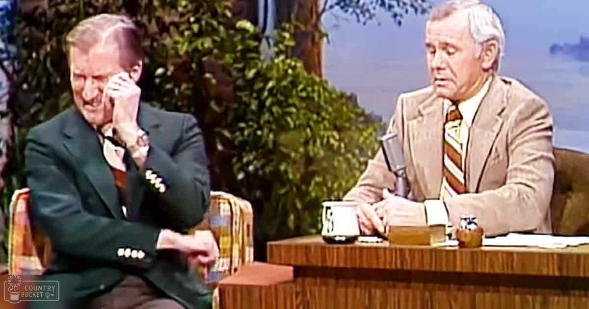 An older adult male singer wearing a dark green suit jacket with white buttons and patterned shirt sits on stage; another older adult male, dressed in a light beige suit jacket over striped collared shirt, speaks into microphone at desk surrounded by plants and outdoor scenery.