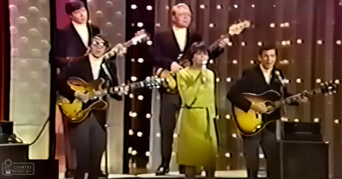A musical performance from the 1960s or early 1970s with a female performer in a bright yellow dress with dark trim, surrounded by musicians playing electric guitars and an acoustic guitar on stage under illuminated circular patterns.