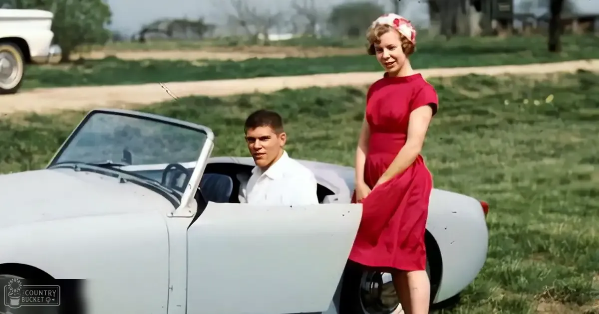 A young couple dressed in 1960s attire stands in an open rural field near a white vintage car.