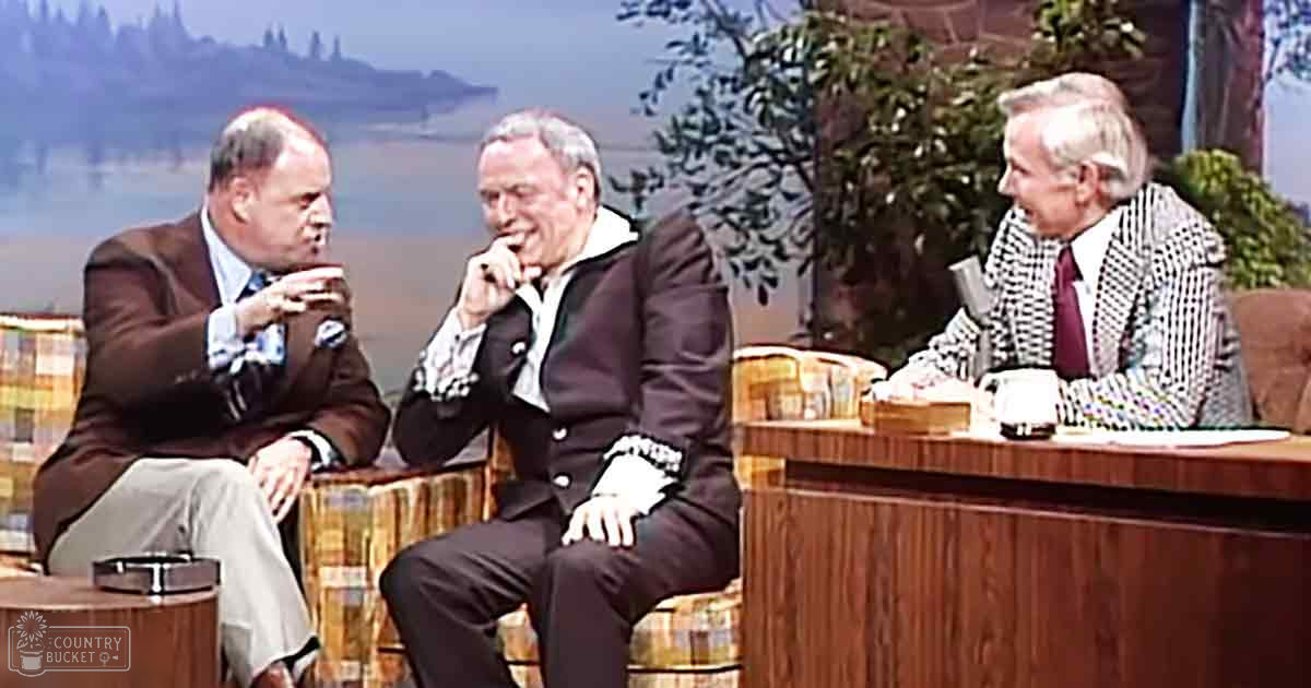 Three adults seated on a couch in what appears to be a television studio set, with wooden furniture and decorative elements like plants. The setting suggests an indoor environment typical of a news or talk show program.