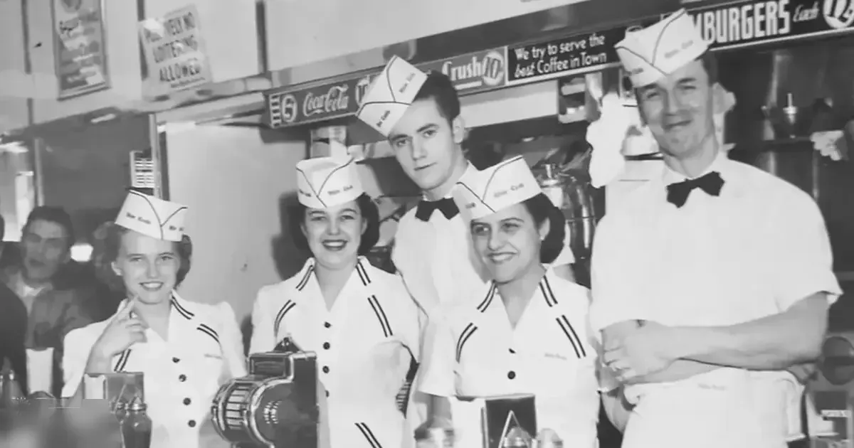 A group of workers in a diner setting; young adult females wear white uniforms with black trim and paper hats, an adult male wears a white shirt with black bow ties, and various diner-related items are visible on the counter.