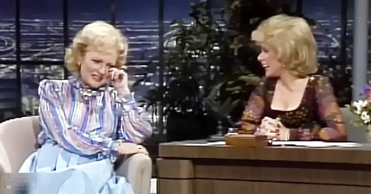 A talk show host in a studio setting with another guest; host wears striped blouse with ruffled collar, guest has dark top with small dots and low-cut neckline, both seated at a wooden desk with urban cityscape background visible through large windows.