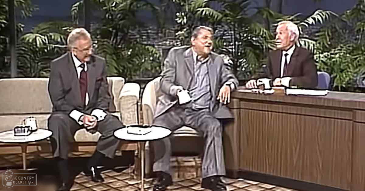 Three men seated in a television talk show studio with armchairs, desks, and potted plants; daytime lighting illuminates the set.
