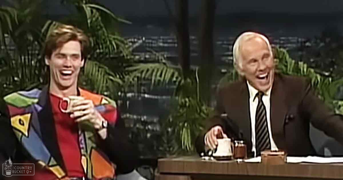 Young adult male in colorful jacket with geometric patterns interacts with older adult male in dark suit and striped tie, set against an indoor studio backdrop with visible greenery and cityscape through windows.
