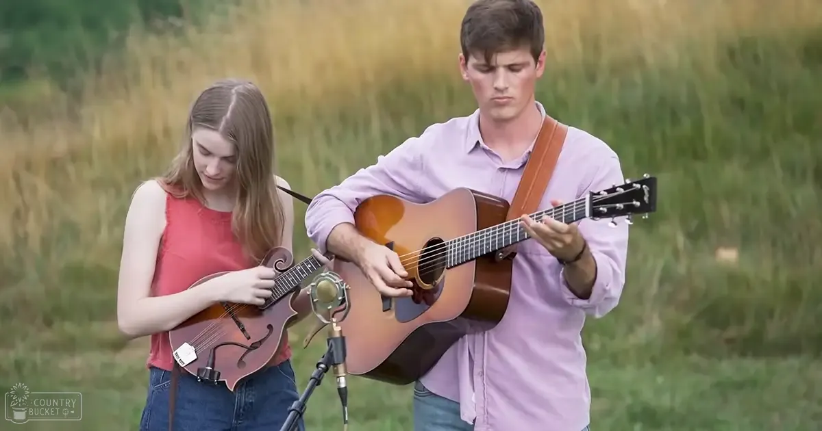 A musical performance with two young adults, one wearing a red sleeveless top and another in a light purple button-up shirt, outdoors amidst green grass under natural daylight.