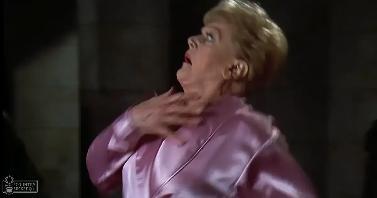 An older adult female wearing a shiny pink satin robe indoors with subdued lighting and neutral-colored walls.