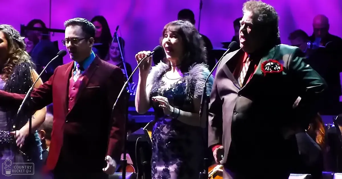 Musical performance in a concert hall; adult male singer wearing glasses and a red vest, female performer holding a microphone in a black mid-20th century dress with lace detailing.