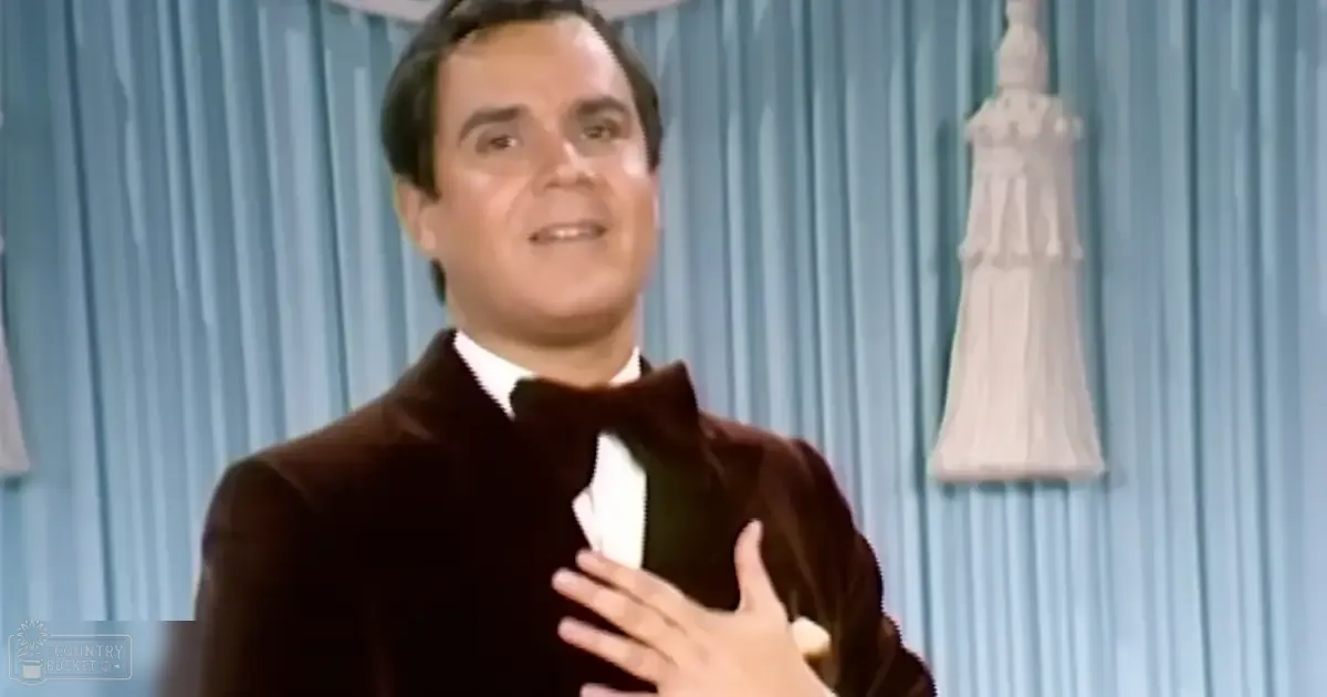 A middle-aged adult male in a tailored dark brown velvet jacket and white shirt, standing indoors with light blue curtains and a decorative sculpture behind him.