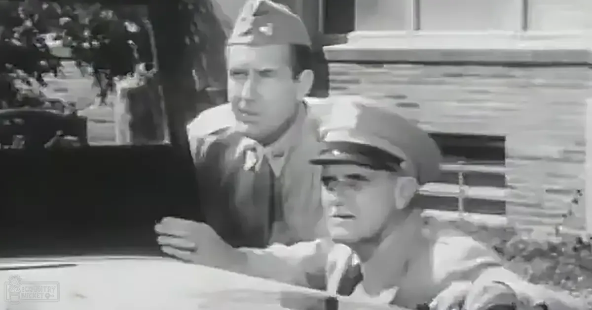 Adult male in military uniform with peaked cap and tie stands near seated young adult male also in similar attire; outdoor scene with trees, structures possibly buildings, daytime, greenery suggesting summer or spring season.