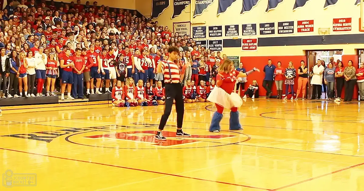 A dance performance in a school gymnasium with young adult dancers, seated spectators, and cheerleaders wearing sports uniforms. The setting includes banners displaying school achievements.
