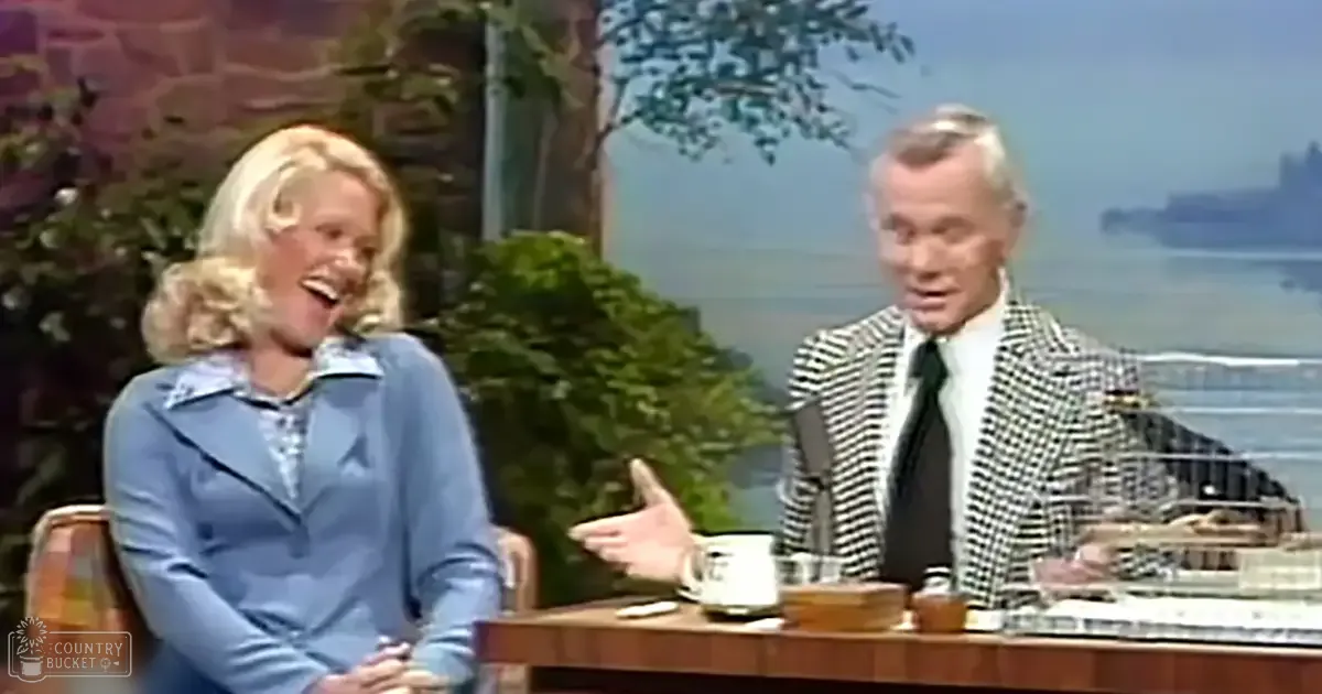 Talk show host and guest in a studio designed to resemble an outdoor scene, with trees and water in the background under bright, even lighting.