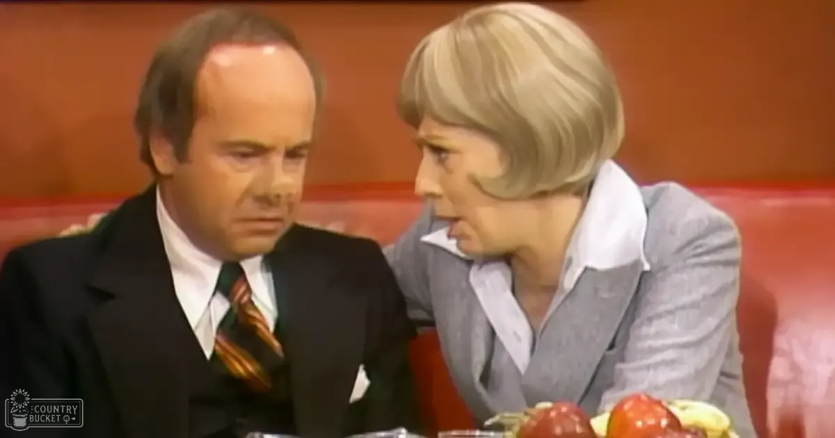 Indoor studio scene resembling a mid-20th century talk show set with two adults in formal attire discussing over food items on a table.