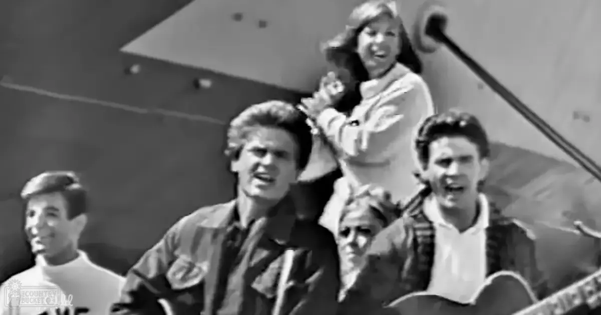 A mid-20th-century American musical performance scene with a young adult male holding an acoustic guitar in the foreground and others in period-appropriate attire, set in what appears to be an aircraft hangar.