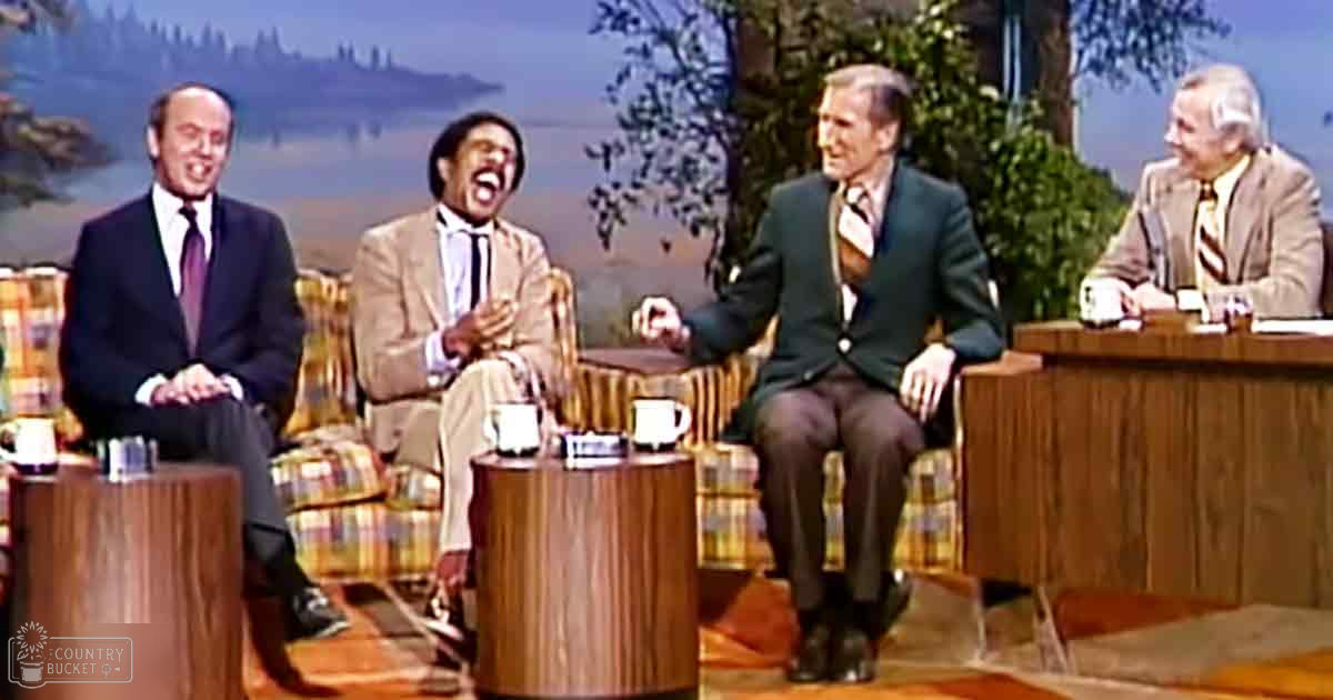 Talk show set with four men seated in a studio environment with earthy tones of furniture against an outdoor backdrop with trees and hills.