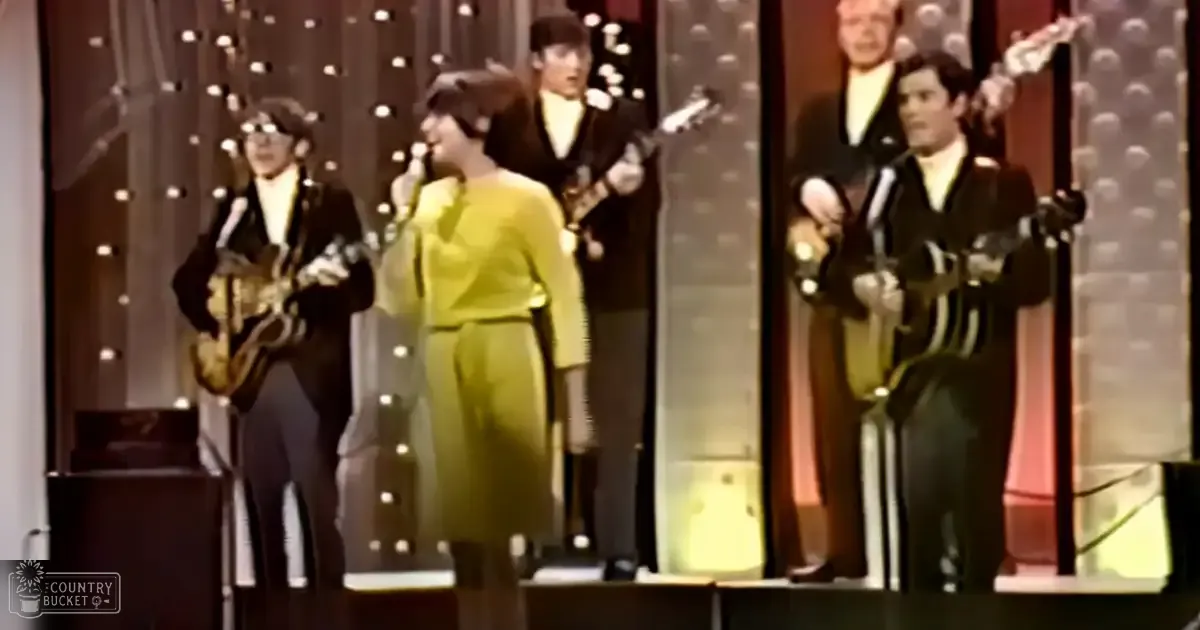 Musical performance scene from past era in studio setting with adult males wearing dark suits holding electric guitars; bright yellow outfit visible.