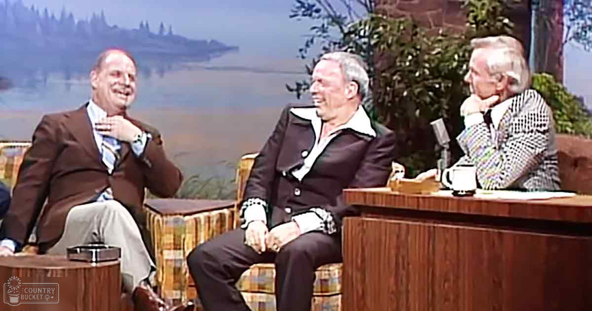 Talk show set with three individuals against an outdoor scenic backdrop with trees and water, suggesting spring or summer daylight.