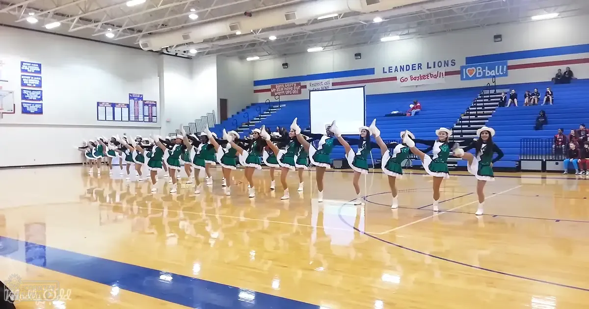 Teenage cheerleaders performing in an indoor school gymnasium with green uniforms, white pom-poms, and a polished wooden floor.