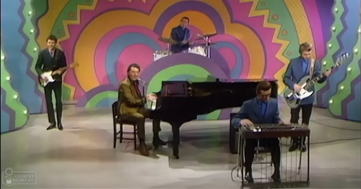 A musical performance with multiple adult males dressed in dark suits playing electric guitars and a piano, set against a vibrant psychedelic backdrop suggesting the 1960s or 1970s.