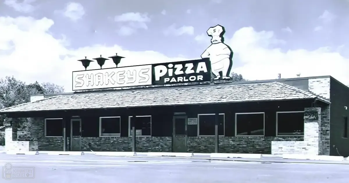 Urban Shakey's Pizza Parlor with chef statue, stone facade, decorative lights, greenery in background, daytime scene.
