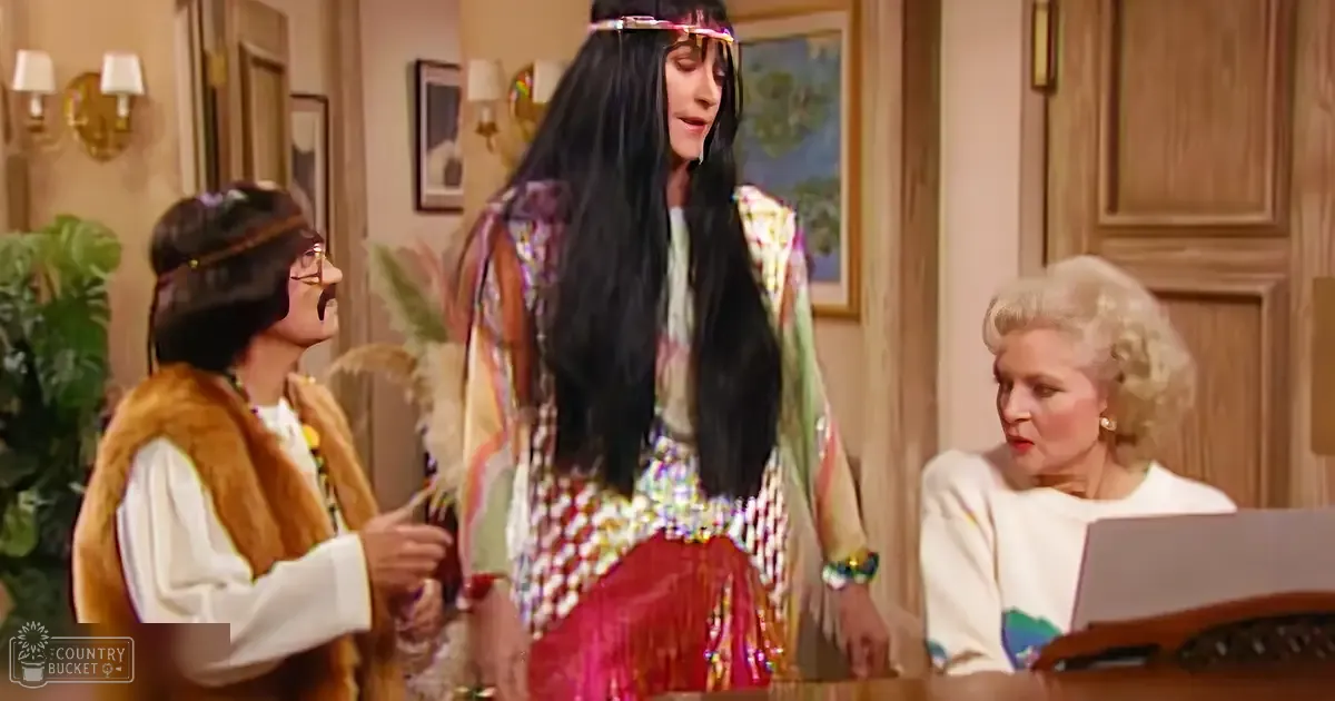 A woman dressed in a colorful patterned dress with long black hair and beads and feathers on her headdress sits indoors, possibly in a home setting.