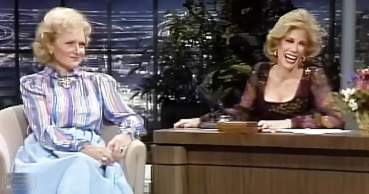 A talk show host and guest seated indoors, with a cityscape visible through large windows at dusk.