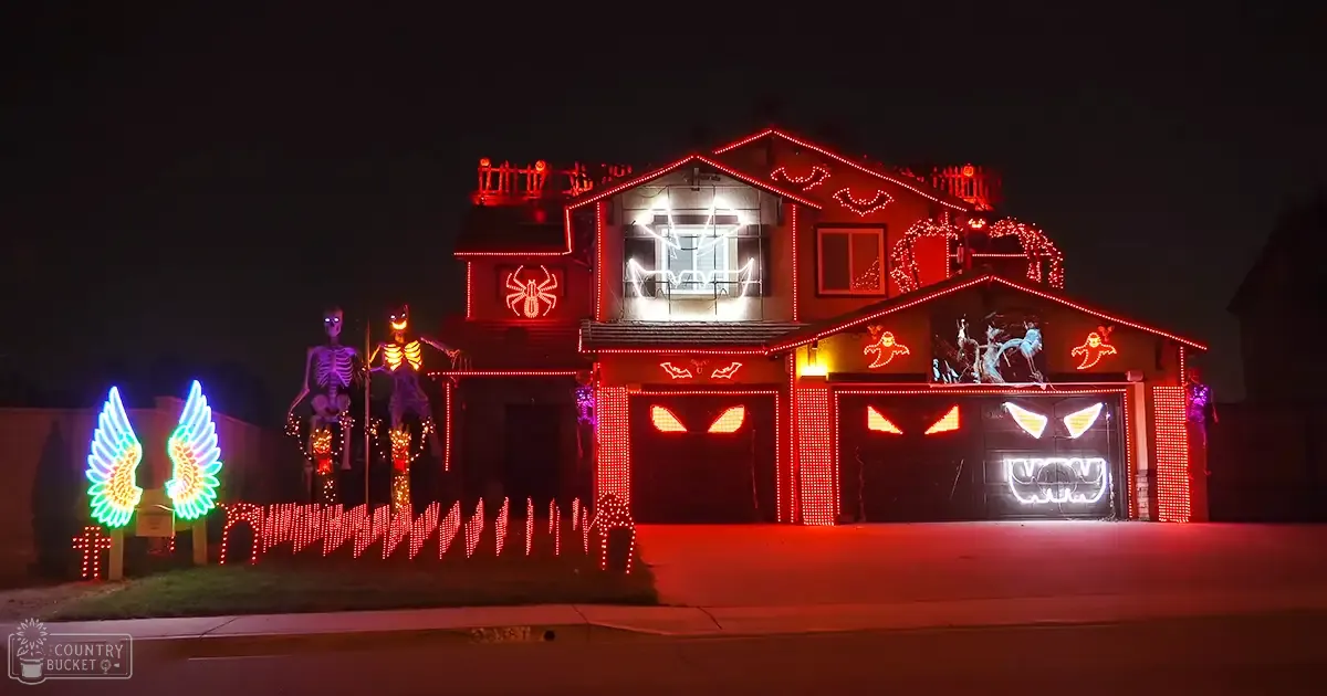 Halloween-themed house decoration scene at night with illuminated skeletons, glowing wings, spider webs, bats, red lights creating a dramatic effect in suburban neighborhood.