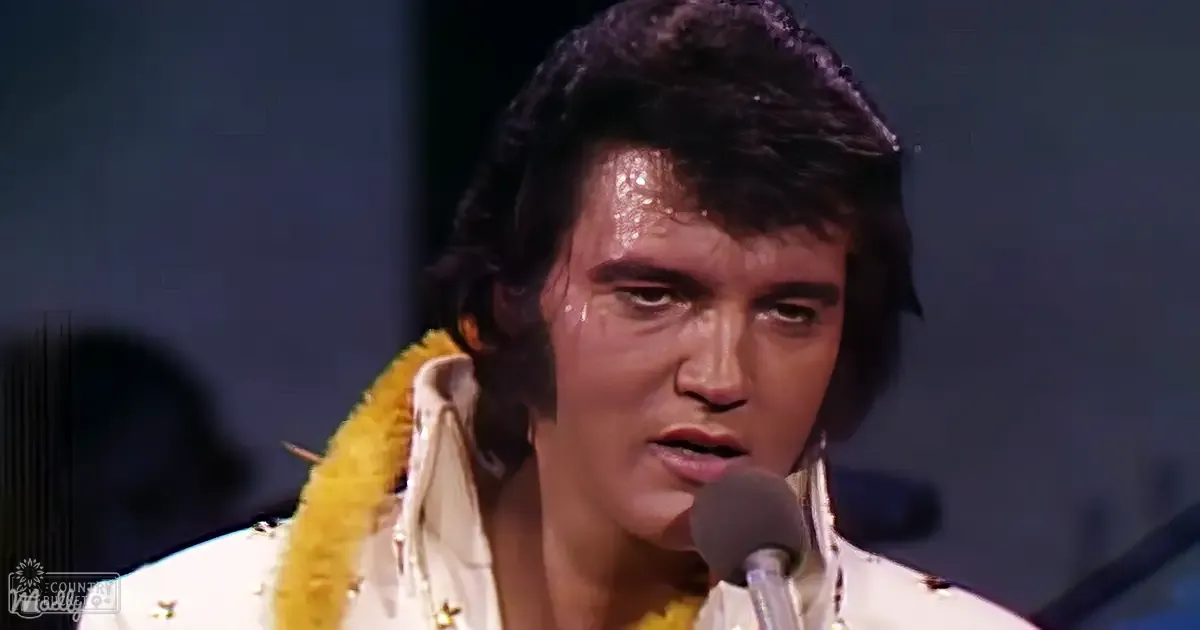 An adult male performer wearing a white, collared, form-fitting jacket adorned with gold fringe and stars, set against a dark stage backdrop with an indoor microphone close by. The setting suggests a recorded performance.