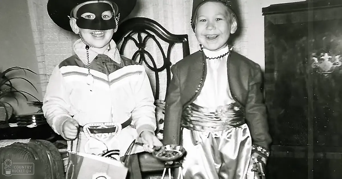 Two children in a home setting with one wearing a white and brown jacket and the other dressed in a white dress with dark jacket, surrounded by furniture like a wooden chair and a potted plant.