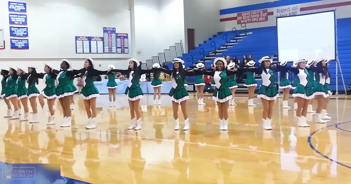 Young female cheerleaders in a modern green and white uniform with ruffles, V-neck tops, and white boots, practicing or performing in an indoor school gymnasium.