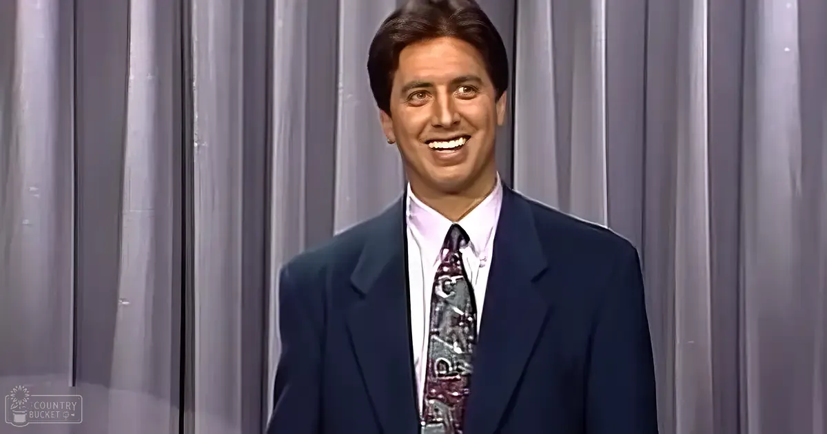 An adult male dressed in a dark blue suit jacket and white dress shirt on stage with gray curtains behind him, likely from the 1980s or early 1990s style.
