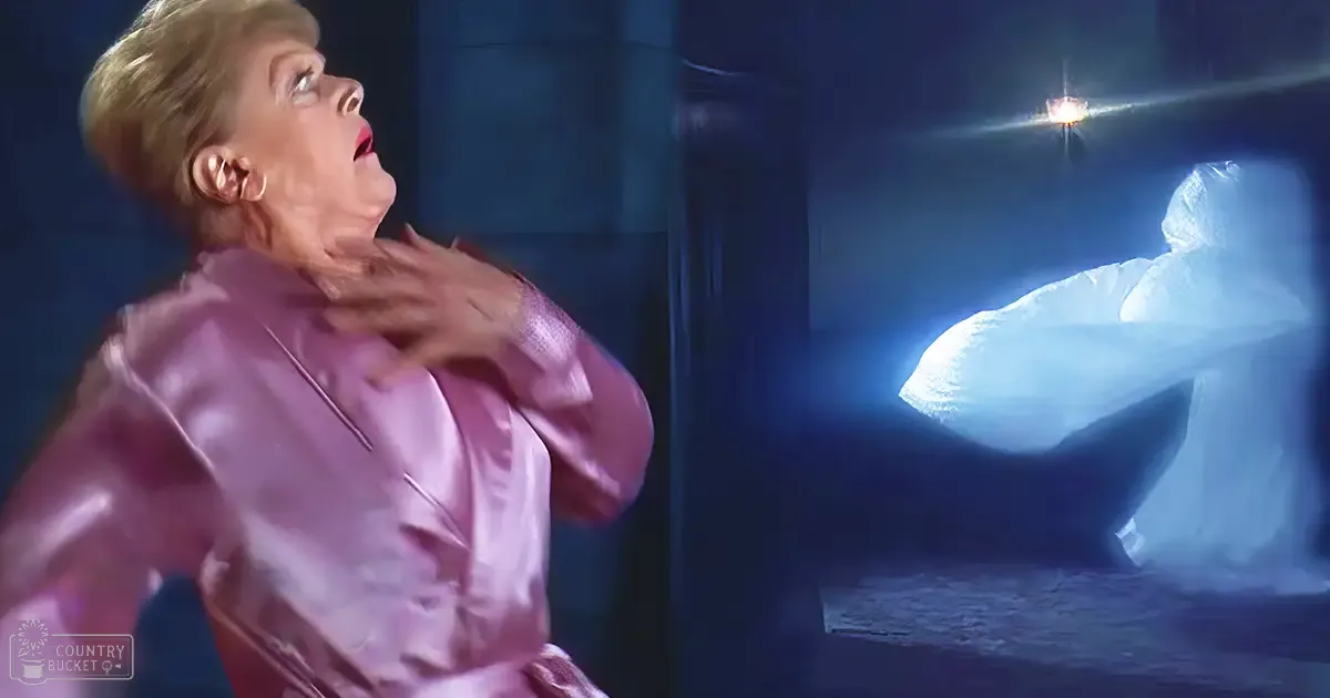 A middle-aged woman wearing a shiny pink robe indoors with dim lighting and a dark ambiance.