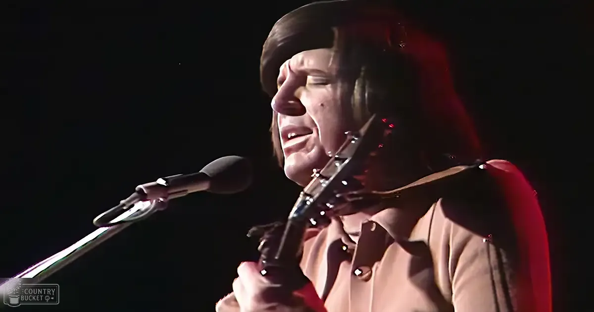 A middle-aged male singer wearing a light-colored button-up shirt performs on stage while holding an acoustic guitar.