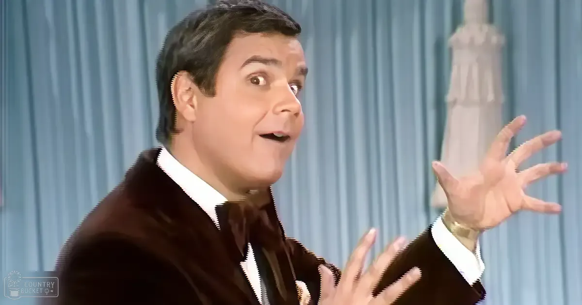 A middle-aged adult male in a dark brown suit jacket and white shirt on an indoor stage with blue curtains, possibly from the mid-20th century.