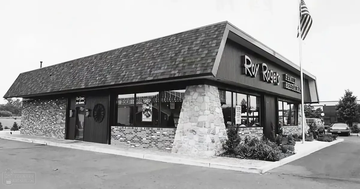 Roy Rogers Family Restaurant building with signage in an urban area, stone pillar, brick facade, windows displaying interior views, American flag on a pole, paved parking lot, shrubs and landscaping, daytime or early evening lighting conditions.