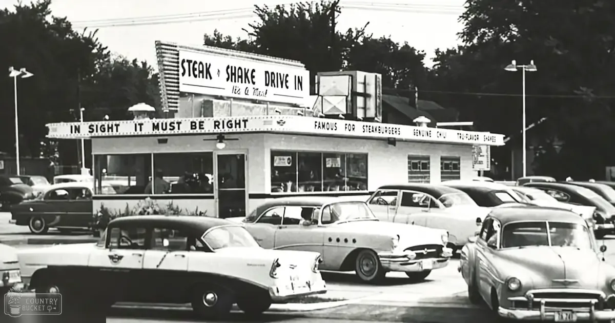 Vintage cars parked at Steak 'n Shake Drive-In in an outdoor parking lot with trees and utility poles, late spring or summer weather, clear daylight conditions, black-and-white historical photograph