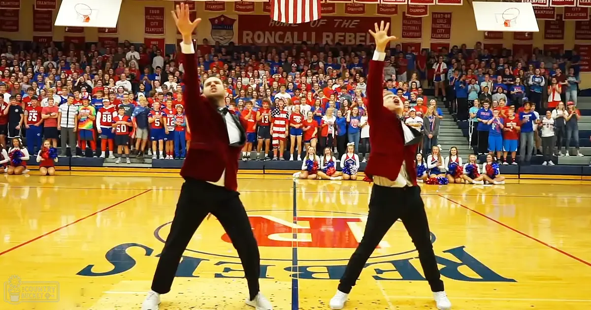 Two young male dancers in red blazers and white shirts performing a routine on stage at the Roncalli Rebels gymnasium with an audience of children and teens watching. The event takes place indoors under bright lighting, surrounded by school banners and flags.