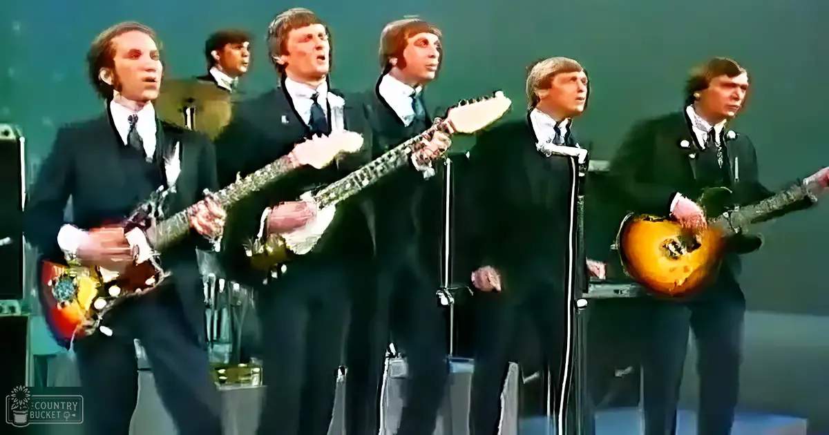 A musical performance scene from the 1960s with young adult male performers in dark suits, holding electric guitars and singing into microphones. The stage backdrop is dark green, with amplifiers and wood-accented guitars visible.