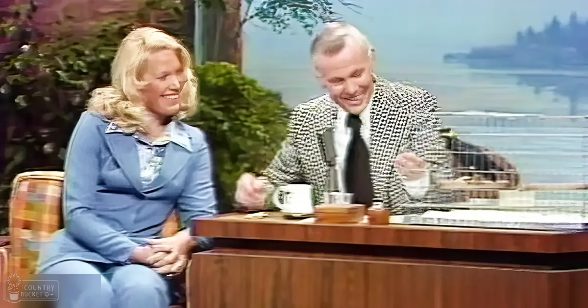 A talk show host and guest are seated at a wooden desk against a scenic outdoor backdrop, likely during daytime with soft natural light.