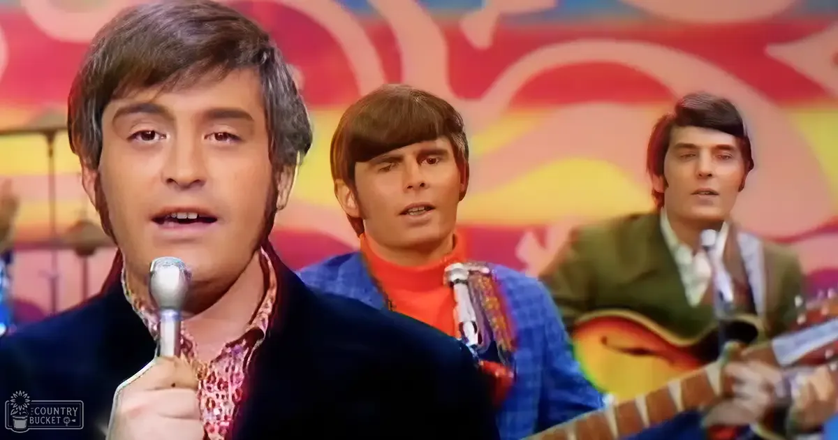 A young adult male singer wearing a dark blue velvet jacket over a patterned shirt with a paisley design and a high collar performs on stage in front of an abstract backdrop with reds, yellows, and blues.