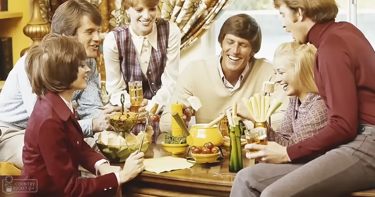 A group of adults gathered indoors, possibly in a home setting with warm lighting. A wooden table holds various items including food and drinks.