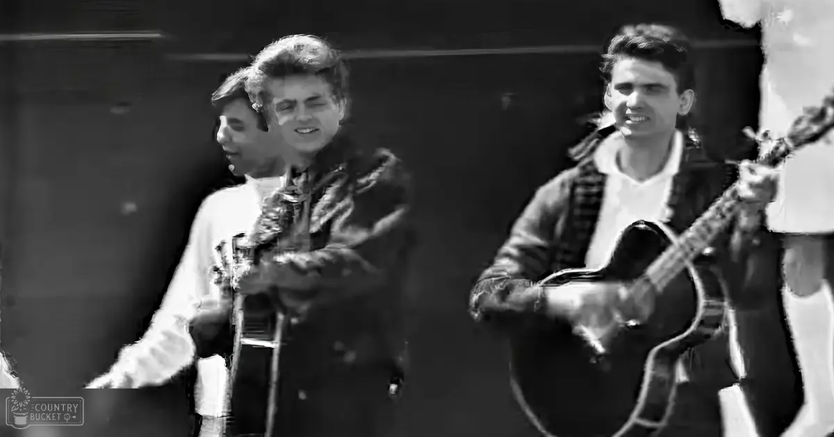 A mid-20th century musical performance scene with young adult males in dark coats and white shirts, one holding a microphone stand while another wears a light-striped skirt or pants and the third holds an acoustic guitar against a backdrop of stage lighting.