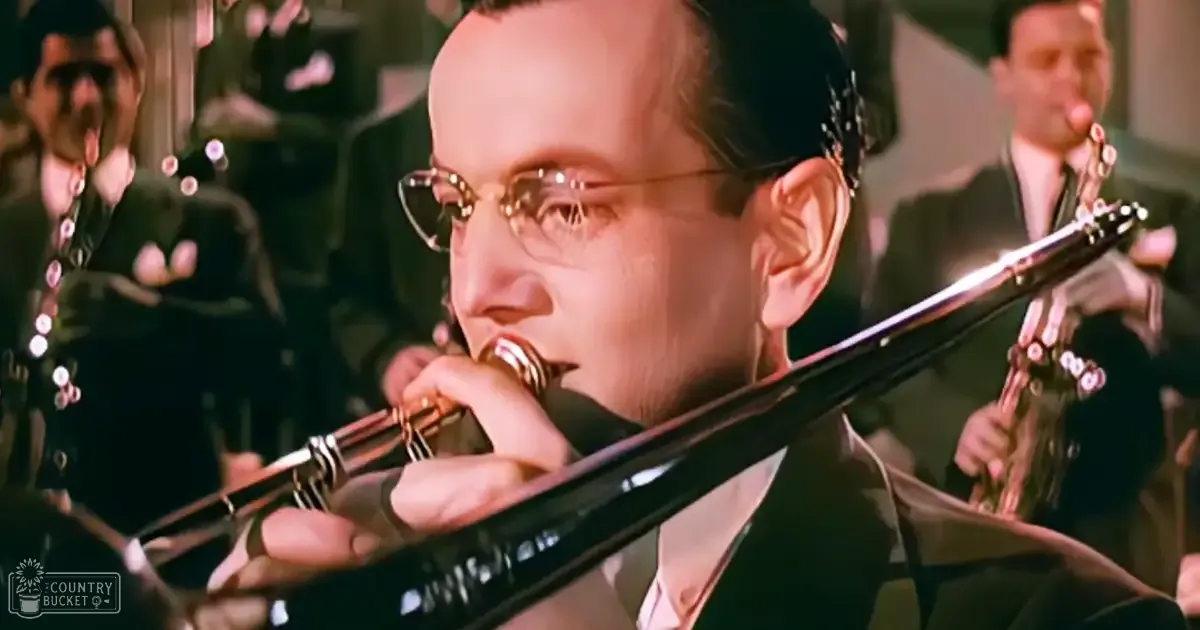 An adult male musician playing a clarinet in an orchestra setting with other musicians visible in dark uniforms and attire, under bright stage lighting.