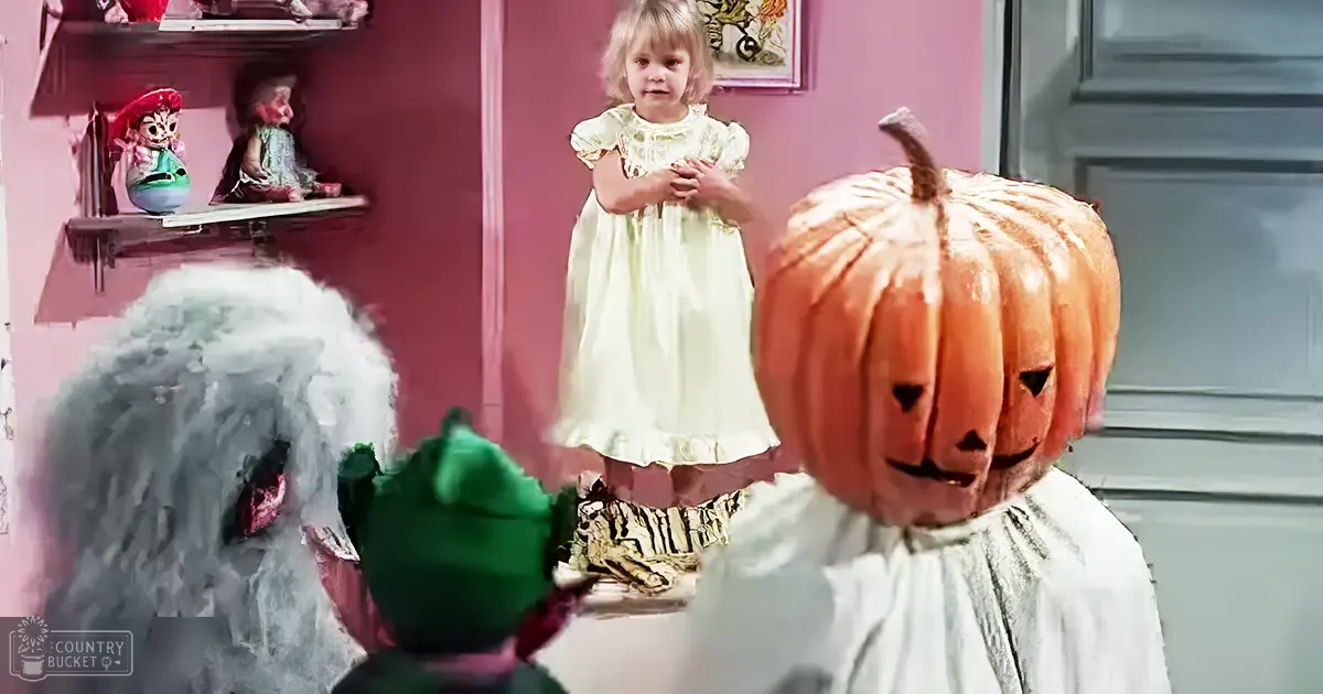A group of toddlers dressed in vintage-style white dresses surrounded by Halloween-themed decorations, suggesting a festive setting.
