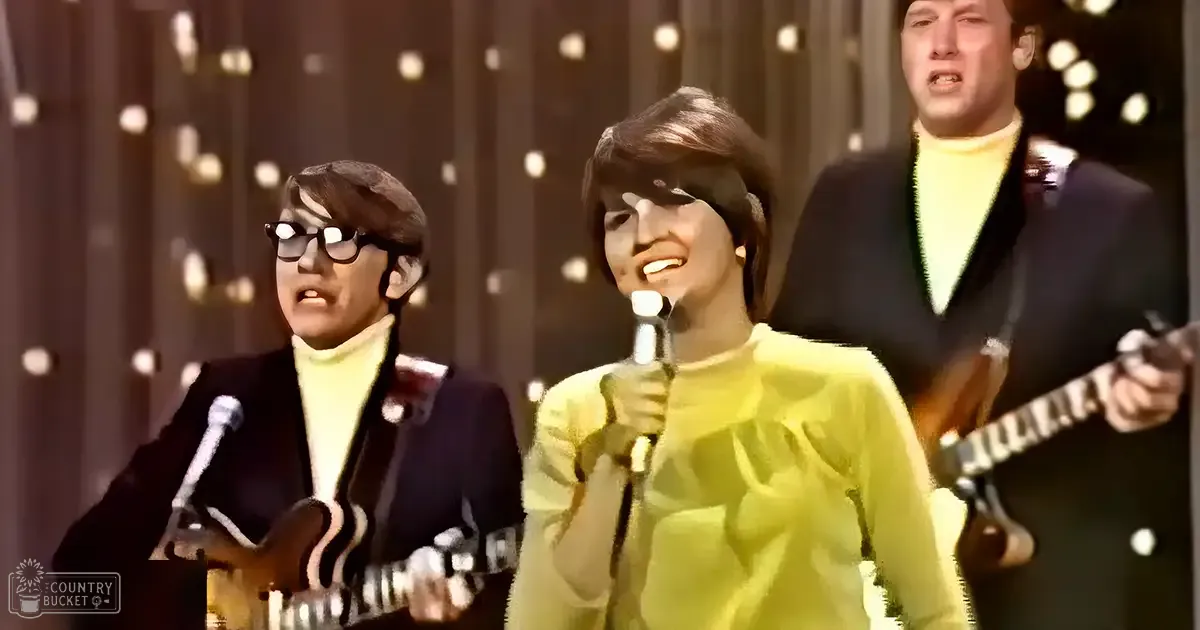 A musical performance scene with young adult males in dark blazers and a young adult female in a light green sweater, set against an indoor backdrop of vertical lines and small circular lights.