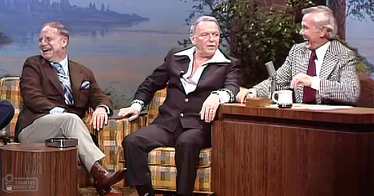 Talk show set with three men, scenic backdrop with trees and mountains, daytime lighting.