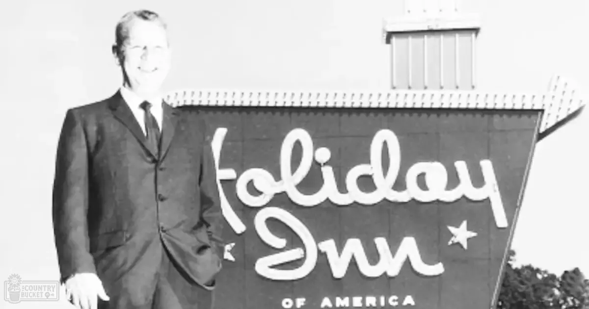 Adult male in dark suit jacket with light shirt and tie stands beside 'Holiday Inn of America' sign at daytime outdoor setting.