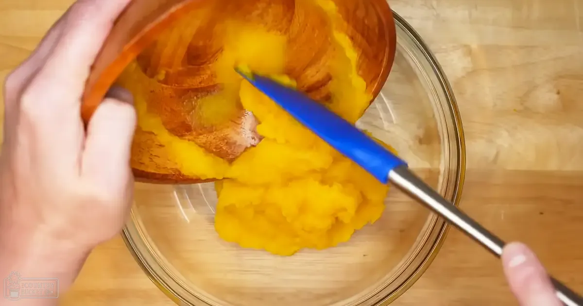 A hand scooping out pumpkin flesh with a knife into a bowl in a kitchen setting, suggesting preparation for cooking or baking.