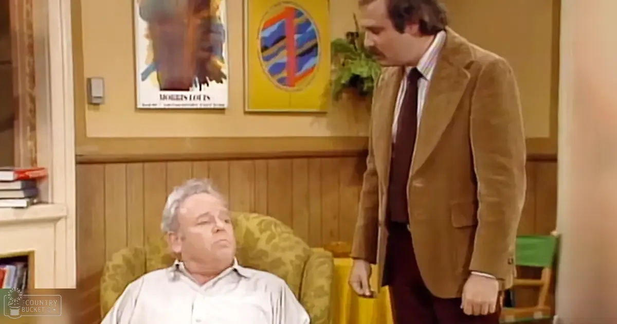 A middle-aged man in a mid-20th-century style button-up shirt sits while another middle-aged male stands wearing a brown corduroy blazer and striped shirt, surrounded by vintage furniture and decor.
